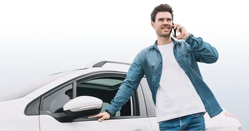 Vyras su džinsiniais marškiniais kalba telefonu atsirėmęs į baltą automobilį